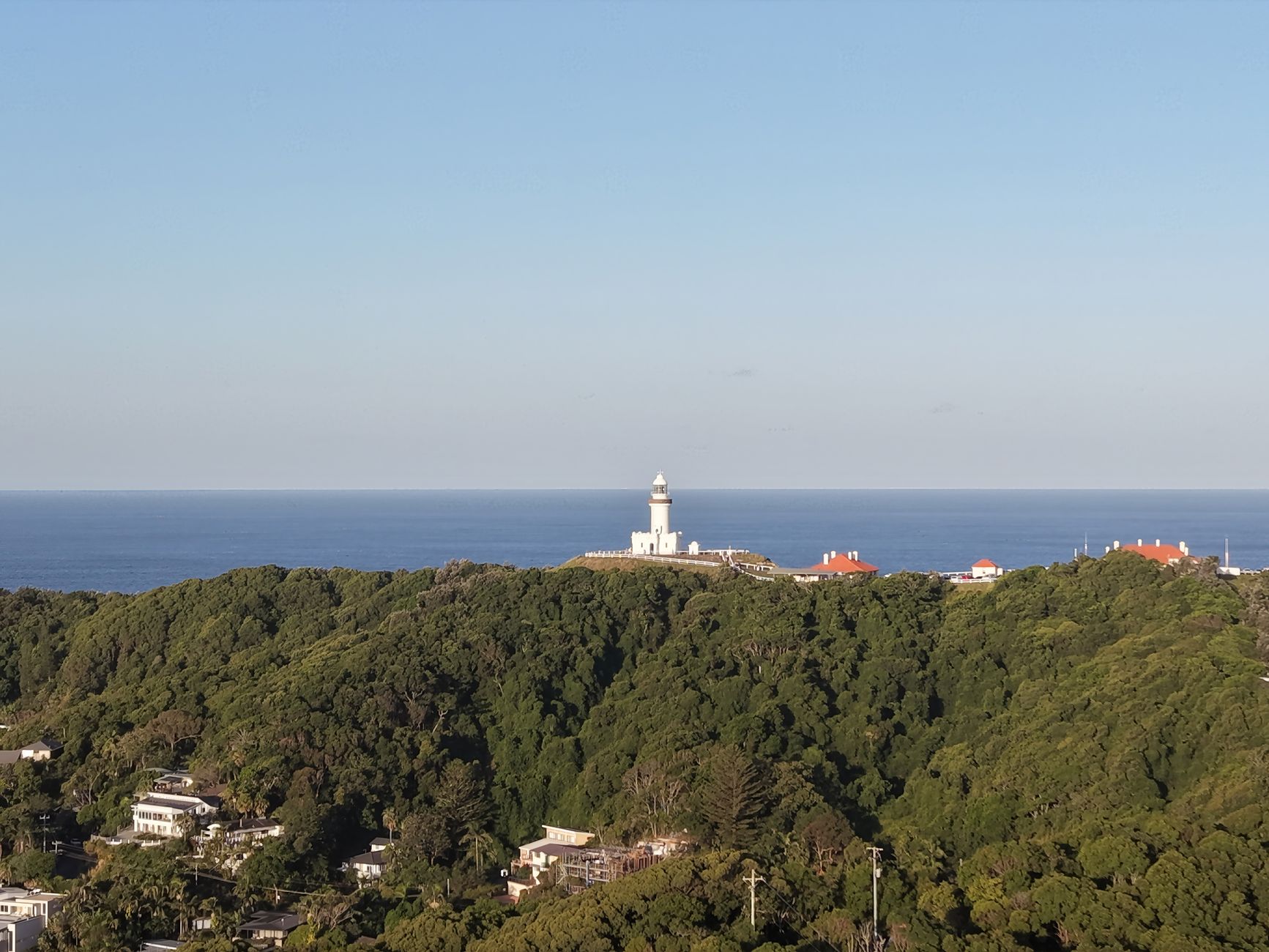 Cape Byron Lighthouse seen from a distance, surrounded by vegetation, with the ocean in the background