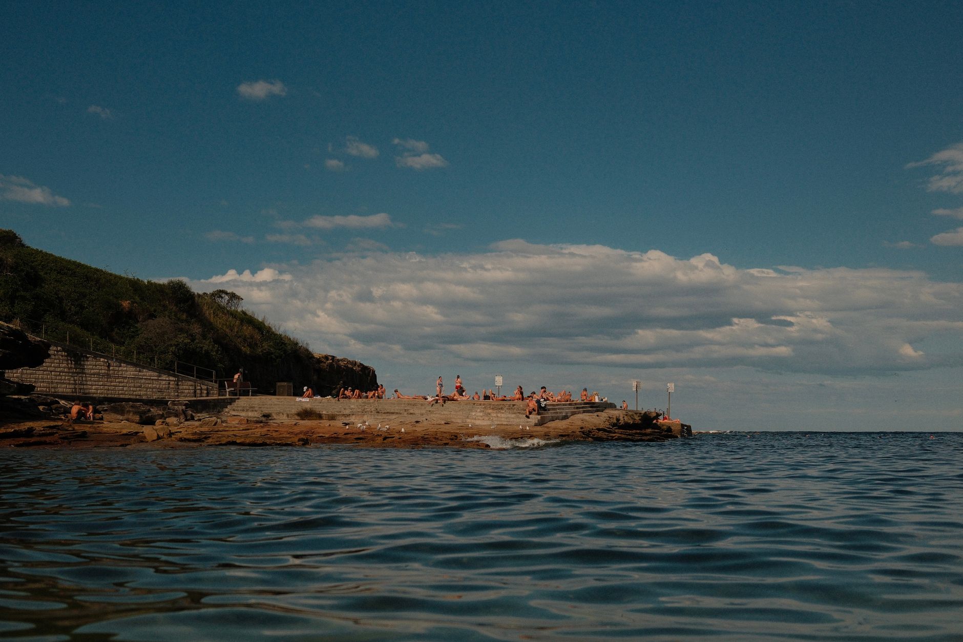 People relaxing on a rocky seaside formation in Sydney, with a green cliff in the background