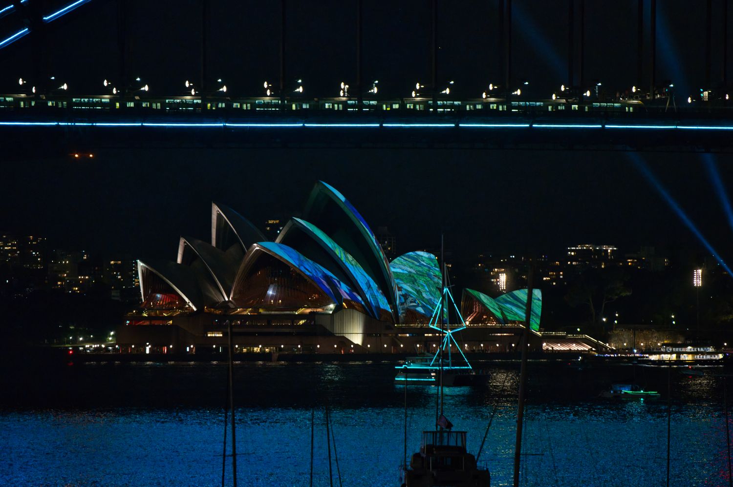 Sydney Opera House lit up in blue and green tones during Vivid Sydney, with the Harbour Bridge also lit up in the background
